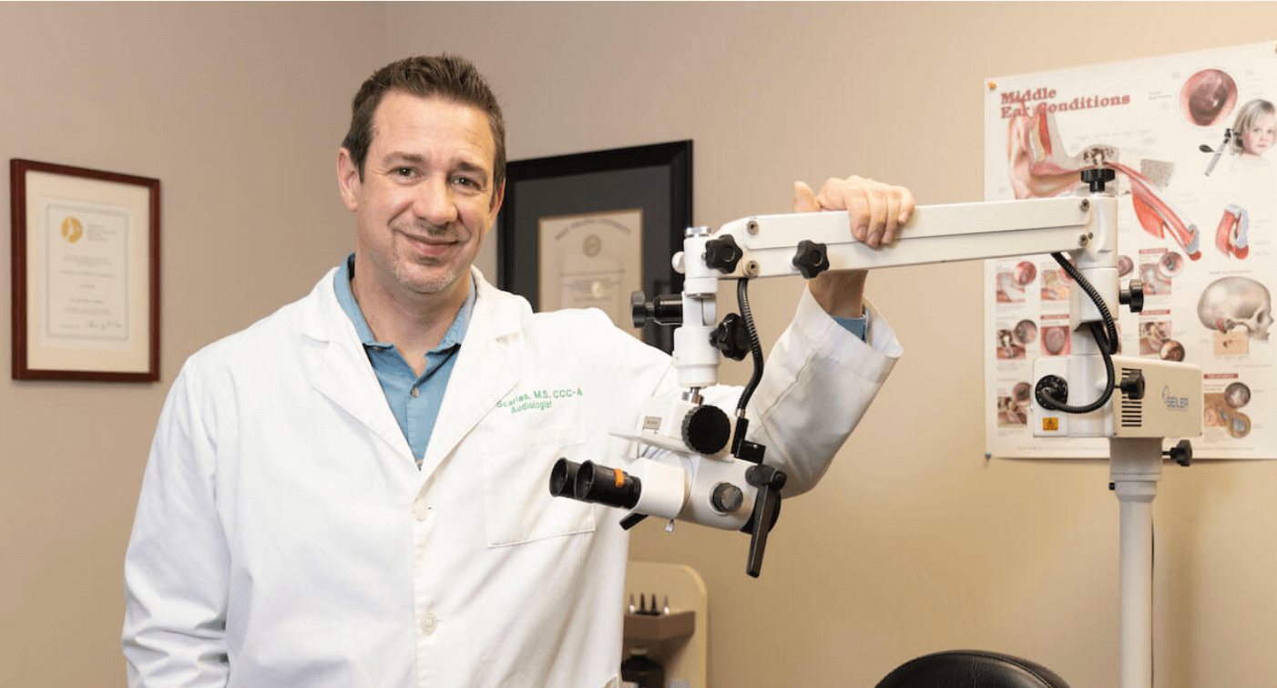 John Scarlas in a white lab coat, standing beside an ear examination microscope in his exam room