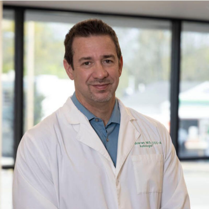 John Scarlas, audiologist, smiling in a white lab coat over a blue shirt